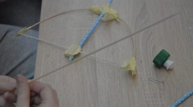 Zdjęcie przedstawiające prototyp łuku strzelniczego. Wykonany jest z wykałaczek, słomki i sznurka. Obok leży zakrętka. Zdjęcie: MBP.