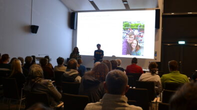 Publiczność siedzi na sali i słucha prezentacji prowadzonej przez dwie osoby stojące przed dużym ekranem. Jedna z osób trzyma mikrofon i zwraca się do uczestników spotkania. Na ekranie wyświetlony jest slajd z tekstem oraz fotografią pary stojącej przy rowerach. Wydarzenie odbywa się w sali audytoryjnej wyposażonej w rzędy krzeseł.