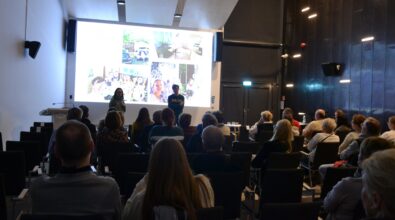 Uczestnicy siedzą na widowni i słuchają prezentacji prowadzonej przez dwie osoby stojące przed dużym ekranem. Jedna z osób mówi do publiczności, trzymając mikrofon. Na ekranie wyświetlony jest slajd z kilkoma fotografiami przedstawiającymi różne momenty z podróży lub spotkań.