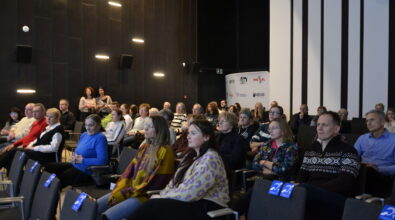 Publiczność siedzi w rzędach krzeseł i uważnie słucha trwającej prezentacji. Uczestnicy patrzą w stronę sceny i ekranu projekcyjnego. Wśród widowni znajdują się osoby w różnym wieku, siedzące obok siebie w sali audytoryjnej. Na niektórych krzesłach umieszczone są oznaczenia z symbolem pętli indukcyjnej.