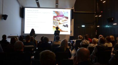 Publiczność siedzi na widowni i słucha prezentacji prowadzonej przez dwie osoby stojące przed ekranem projekcyjnym. Jedna z osób trzyma mikrofon i mówi do uczestników spotkania. Na ekranie wyświetlony jest slajd z tytułem „Kuchnie Toskanii” oraz fotografią potraw. Wydarzenie odbywa się w sali audytoryjnej wyposażonej w rzędy krzeseł.