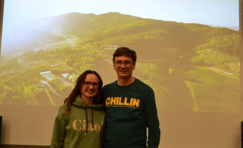 Karolina Iskierka i Paweł Sierszeń stoją przed dużym ekranem projekcyjnym z wyświetlonym slajdem przedstawiającym krajobraz z winnicą. Oboje uśmiechają się do aparatu. Za nimi widoczny jest tytuł prezentacji „Jak adoptowaliśmy kawałek winnicy? – Palagina”. Prelegenci pozują do zdjęcia na scenie w