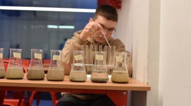 Na zdjęciu widać siedem zlewek z roztworem wody oraz gleby. W tle widać nastolatka trzymającego w dłoni pipetę. Przed nastolatkiem stoi również osiem próbówek. Zdjęcie własnością MBP Piotrków.