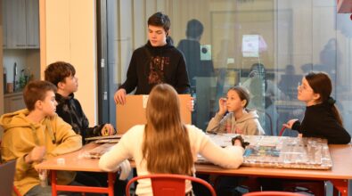 Zdjęcie przedstawia piątkę dzieci i nastolatka. Znajdują się oni przy dużym stole, na którym leży duży kawałek tektury. Dzieci siedzą dookoła stołu. Nastolatek stoi, mówi coś do reszty, która go uważnie słucha. W dłoni trzyma kawałek tektury. Za nimi znajduje się okno z widokiem na pomieszczenie, do którego przejście widać w tle po lewej stronie zdjęcia. Zdjęcie własnością MBP Piotrków.