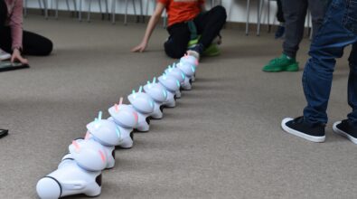 Zdjęcie przedstawia ustawione w rzędzie niewielkie białe roboty. Za nimi siedzi chłopiec. Zdjęcie wykonano podczas zajęć, dzieci prawdopodobnie przygotowują roboty do wykonania sekwencji wyznaczonych ruchów. Zdjęcie: MBP.