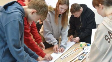 Grupa młodzieży stoi wokół stołu i wspólnie pracuje nad papierową konstrukcją, przyklejając elementy taśmą i dopasowując detale. Uczestnicy są skupieni na zadaniu, a na blacie widać papierowe rulony, klej, taśmę i przybory piśmiennicze. Zdjęcie przedstawia zespołową pracę młodzieży podczas konkursu konstrukcyjnego w bibliotece.