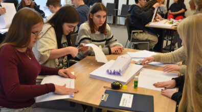 Przy jednym stole siedzi grupa młodych osób, które wycinają, składają i przygotowują papierowe elementy konstrukcji. Uczestnicy są skupieni na pracy, a na blacie leżą kartki papieru, nożyczki, klej oraz taśma miernicza. Zdjęcie przedstawia zespołową pracę młodzieży podczas konkursu konstrukcyjnego w przestrzeni bibliotecznej.