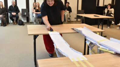 Uśmiechnięta kobieta pochyla się nad stołem i puszcza mały samochodzik po papierowej rampie wykonanej z kartek i taśmy. Konstrukcja łączy dwa stoły i jest elementem zadania konkursowego, a w tle na krzesłach siedzą uczestnicy i obserwują przebieg próby. Zdjęcie przedstawia moment testowania papierowej konstrukcji podczas drużynowego konkursu w przestrzeni bibliotecznej.