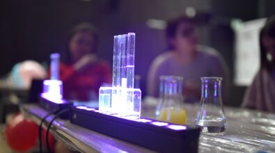 Zdjęcie przedstawia lampę ultrafiolet, na której stoją próbówki. W nic znajdują się różne ciecze, z których jedna świeci jasnobiałym światłem. W tle widać kolby z cieczami, a dalej uczestników warsztatów. Zdjęcie: MBP.