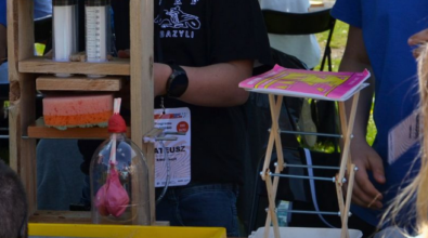 Na zdjęciu widać konstrukcję wykonaną z desek, strzykawek oraz gąbki. Po prawej stronie znajduje się składany stojak wykonany z drewna i metalu, na którym utrzymuje się zeszyt. Chłopiec stojący z tyłu ma na szyi identyfikator z logo wydarzenia. Zdjęcie: MBP.