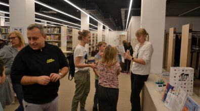 Grupa osób stojących przy ladzie w nowoczesnej przestrzeni bibliotecznej między regałami z książkami, które rozmawiają i biorą udział w wspólnym działaniu. Wnętrze jest jasno oświetlone, a uczestnicy skupieni na wykonywanych czynnościach, co sugeruje spotkanie lub warsztaty odbywające się w bibliotece.