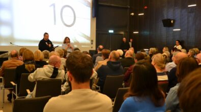 Na zdjęciu widoczna jest licznie zgromadzona publiczność w sali Mediateki 800-lecia, siedząca tyłem do obiektywu i skierowana w stronę sceny z ekranem projekcyjnym. Uczestnicy spotkania biorą udział w dyskusji, a jedna z osób na widowni podnosi rękę, sygnalizując chęć zabrania głosu.