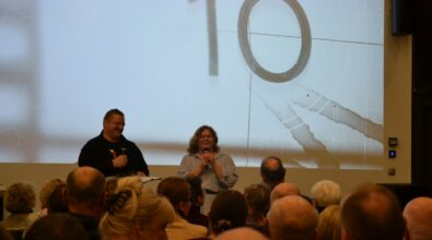 Na zdjęciu widoczni są prowadzący spotkanie stojący przed ekranem projekcyjnym z wyświetloną grafiką, zwróceni do licznie zgromadzonej publiczności w sali Mediateki 800-lecia. Uczestnicy siedzą na widowni i uważnie słuchają wystąpienia, które ma formę rozmowy i komentarzy do prezentowanych treści.