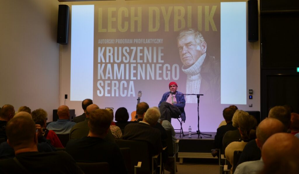 Lech Dyblik siedzi na scenie, z gitarą i projekcją wizualną w tle, przed nim zgromadzona publiczność.