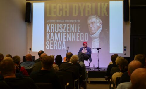 Lech Dyblik siedzi na scenie, z gitarą i projekcją wizualną w tle, przed nim zgromadzona publiczność.
