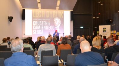 Lech Dyblik siedzi na scenie, obok niego gitara na stojaku i projekcja wizualna w tle, przed nim zgromadzona publiczność.