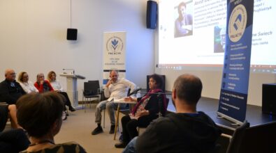 Zdjęcie przedstawia spotkanie dyskusyjne w sali Mediateki, w którym uczestniczy grupa dorosłych osób siedzących w półokręgu na krzesłach. W centrum widoczna jest kobieta mówiąca do mikrofonu, obok niej siedzi mężczyzna, a rozmowa ma charakter panelu lub otwartej debaty. W tle znajduje się ekran z prezentacją multimedialną zawierającą zdjęcia i tekst dotyczący tematyki psychologicznej i społecznej. Po bokach sali ustawione są roll-upy organizacji społecznej z hasłami dotyczącymi wsparcia, integracji i pomocy wzajemnej. Uczestnicy są zwróceni w stronę prelegentów, a atmosfera spotkania ma charakter uważnej, zaangażowanej rozmowy.