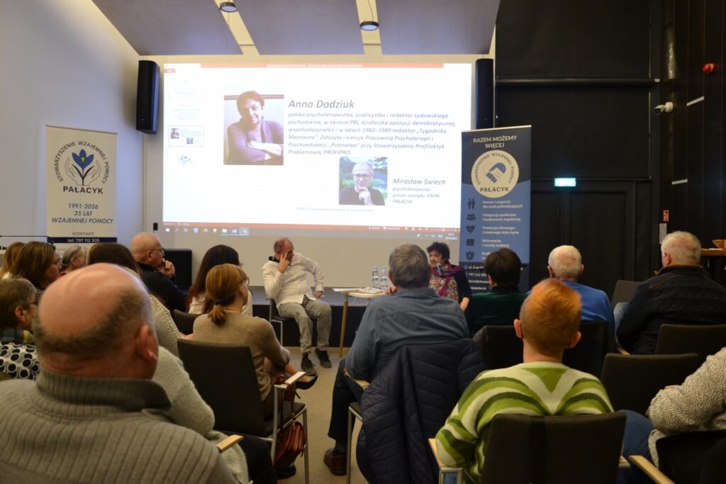 Zdjęcie przedstawia spotkanie otwarte w sali Mediateki, w którym uczestniczy liczna grupa dorosłych osób siedzących na krzesłach i zwróconych w stronę prelegentów. Na pierwszym planie widoczna jest publiczność, a w centralnej części sali kobieta i mężczyzna prowadzący rozmowę, siedzący przy niewielkim stoliku. W tle znajduje się duży ekran z prezentacją multimedialną zawierającą zdjęcia oraz informacje biograficzne dotyczące Anny Dodziuk i Mirosława Świecha. Po obu stronach ekranu ustawione są roll-upy Stowarzyszenia Wzajemnej Pomocy Palacyk z hasłami dotyczącymi wsparcia, integracji społecznej i działań pomocowych. Atmosfera spotkania jest skupiona i sprzyja uważnemu słuchaniu oraz refleksji.