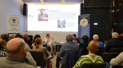 Zdjęcie przedstawia spotkanie otwarte w sali Mediateki, w którym uczestniczy liczna grupa dorosłych osób siedzących na krzesłach i zwróconych w stronę prelegentów. Na pierwszym planie widoczna jest publiczność, a w centralnej części sali kobieta i mężczyzna prowadzący rozmowę, siedzący przy niewielkim stoliku. W tle znajduje się duży ekran z prezentacją multimedialną zawierającą zdjęcia oraz informacje biograficzne dotyczące Anny Dodziuk i Mirosława Świecha. Po obu stronach ekranu ustawione są roll-upy Stowarzyszenia Wzajemnej Pomocy Palacyk z hasłami dotyczącymi wsparcia, integracji społecznej i działań pomocowych. Atmosfera spotkania jest skupiona i sprzyja uważnemu słuchaniu oraz refleksji.