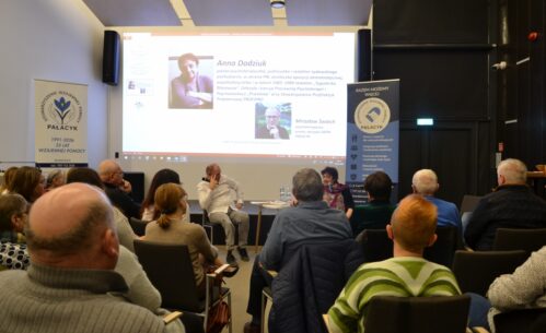 Zdjęcie przedstawia spotkanie otwarte w sali Mediateki, w którym uczestniczy liczna grupa dorosłych osób siedzących na krzesłach i zwróconych w stronę prelegentów. Na pierwszym planie widoczna jest publiczność, a w centralnej części sali kobieta i mężczyzna prowadzący rozmowę, siedzący przy niewielkim stoliku. W tle znajduje się duży ekran z prezentacją multimedialną zawierającą zdjęcia oraz informacje biograficzne dotyczące Anny Dodziuk i Mirosława Świecha. Po obu stronach ekranu ustawione są roll-upy Stowarzyszenia Wzajemnej Pomocy Palacyk z hasłami dotyczącymi wsparcia, integracji społecznej i działań pomocowych. Atmosfera spotkania jest skupiona i sprzyja uważnemu słuchaniu oraz refleksji.