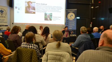 Zdjęcie przedstawia liczne spotkanie publiczne w sali Mediateki, w którym uczestniczy grupa dorosłych osób siedzących na krzesłach i zwróconych w stronę prelegentki prowadzącej dyskusję. Na pierwszym planie widoczna jest publiczność słuchająca wystąpienia, a w centralnej części kobieta gestykulująca podczas wypowiedzi. W tle znajduje się duży ekran z prezentacją multimedialną zawierającą zdjęcia oraz informacje biograficzne dotyczące Anny Dodziuk i Mirosława Świecha. Po prawej stronie sali ustawiony jest roll-up organizacji Palacyk z hasłami dotyczącymi wzajemnej pomocy, integracji społecznej i wsparcia osób potrzebujących. Atmosfera spotkania ma charakter zaangażowanej debaty i sprzyja refleksji oraz wymianie myśli.