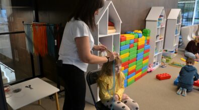 Kobieta stojącą obok siedzącej dziewczynki, która ma zakładane lub poprawiane akcesorium na głowie podczas zajęć w przestrzeni bibliotecznej. W tle znajdują się kolorowe klocki oraz regały z materiałami edukacyjnymi, co wskazuje na warsztaty lub animację dla dzieci odbywającą się w bibliotece.