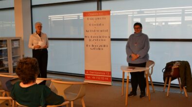 Dwie osoby stojące w sali spotkań przy stolikach, obok roll-upu informacyjnego, podczas wydarzenia organizowanego w przestrzeni bibliotecznej. Uczestnicy zgromadzeni są na sali, a całość sugeruje prelekcję lub spotkanie autorskie odbywające się w bibliotece.