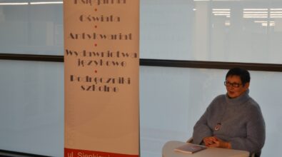 Osoba siedząca przy stoliku w sali spotkań oraz stojący obok roll-up informacyjny z danymi instytucji i ofertą wydarzeń kulturalnych. Wnętrze biblioteki jest jasno oświetlone, a kadr sugeruje fragment prelekcji lub spotkania odbywającego się w przestrzeni Mediateki.