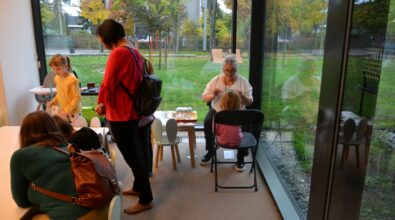 Grupa osób przebywających w przestrzeni bibliotecznej przy dużych oknach z widokiem na zieleń, część siedzi na krzesłach, a część stoi i rozmawia. Uczestnicy spotkania spędzają czas w swobodnej atmosferze, co sugeruje integracyjne wydarzenie lub warsztaty odbywające się w Mediatece.