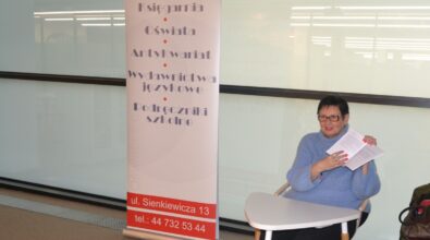 Osoba siedząca przy małym stoliku w sali spotkań, trzymająca kartkę i uśmiechająca się w stronę uczestników. Obok znajduje się roll-up informacyjny z nazwą i danymi instytucji, a całość wskazuje na kameralne spotkanie lub prelekcję odbywającą się w przestrzeni bibliotecznej.