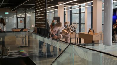 Grupa osób siedzących przy stolikach w nowoczesnej przestrzeni bibliotecznej z przeszklonymi balustradami, uczestniczących we wspólnym spotkaniu. Jasne wnętrze i widoczne regały oraz stoły wskazują na zajęcia lub wydarzenie odbywające się w Mediatece.