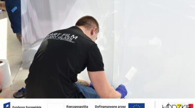 Na zdjęciu widać mężczyznę montującego folię ciekłokrystaliczną na dużej, przeszklonej powierzchni, pracującego w pozycji pochylonej i używającego rękawic ochronnych. Na dole grafiki umieszczone są logotypy Funduszy Europejskich dla Łódzkiego, Rzeczypospolitej Polskiej, Unii Europejskiej oraz Urzędu Marszałkowskiego Województwa Łódzkiego, co wskazuje na realizację zadania współfinansowanego ze środków europejskich.