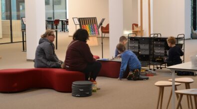 Dzieci bawiące się klockami na dywanie w przestrzeni bibliotecznej, a obok nich siedzących dorosłych opiekunów obserwujących zabawę. W tle znajdują się regały z materiałami edukacyjnymi oraz elementy wyposażenia sali, co wskazuje na zajęcia lub swobodną aktywność dzieci odbywającą się w bibliotece.