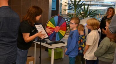 Animatorka prowadzącą zabawę edukacyjną dla dzieci w przestrzeni Mediateki. Kobieta stoi przy stoliku z kolorowym kołem fortuny podzielonym na pola z nazwami zadań i aktywności, a obok znajduje się tabliczka z informacją o godzinach wydarzenia. Przed animatorką stoi grupa dzieci w różnym wieku, uważnie słuchających wyjaśnień i oczekujących na swoją kolej. W tle widoczni są dorośli opiekunowie oraz elementy przestrzeni bibliotecznej z roślinami i przeszklonymi ścianami. Atmosfera spotkania jest dynamiczna i angażująca, sprzyjająca zabawie i nauce.