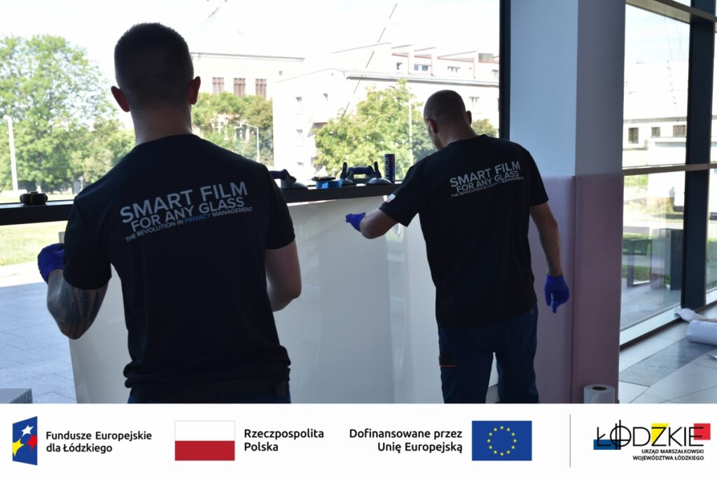 Dwóch mężczyzn montuje inteligentną folię na dużym oknie – działanie w ramach projektu wspieranego przez fundusze europejskie. Na dole zdjęcia widoczne są logotypy instytucji wspierających projekt.