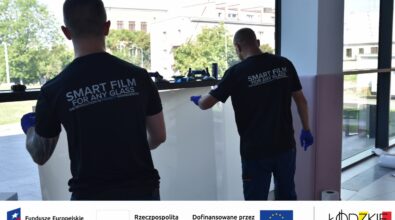 Dwóch mężczyzn montuje inteligentną folię na dużym oknie – działanie w ramach projektu wspieranego przez fundusze europejskie. Na dole zdjęcia widoczne są logotypy instytucji wspierających projekt.