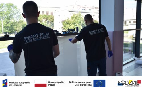 Dwóch mężczyzn montuje inteligentną folię na dużym oknie – działanie w ramach projektu wspieranego przez fundusze europejskie. Na dole zdjęcia widoczne są logotypy instytucji wspierających projekt.