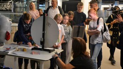 Grupa dzieci i dorosłych zgromadzonych wokół stołu w przestrzeni bibliotecznej, obserwujących pokaz lub doświadczenie prowadzone przez instruktorkę. Uczestnicy z zainteresowaniem przyglądają się prezentowanemu urządzeniu i wykonywanym czynnościom, a całość odbywa się w nowoczesnym, otwartym wnętrzu Mediateki.