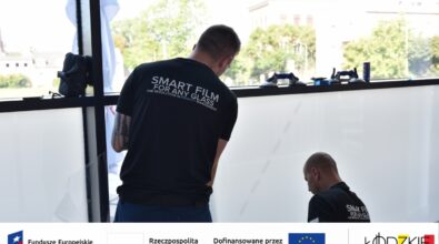 Dwóch mężczyzn montuje inteligentną folię na dużym oknie – działanie w ramach projektu wspieranego przez fundusze europejskie. Na dole zdjęcia widoczne są logotypy instytucji wspierających projekt.