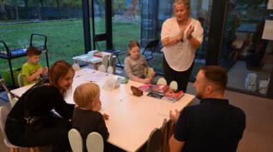 Przy stole siedzi kilkoro dzieci oraz dorosłych, którzy wspólnie uczestniczą w zajęciach plastycznych. Na blacie znajdują się kartki papieru, kredki i inne materiały do pracy twórczej. Jedna z kobiet stoi obok stołu i klaszcze w dłonie, zachęcając dzieci do działania, a w tle przez duże okna widać zielony teren na zewnątrz.