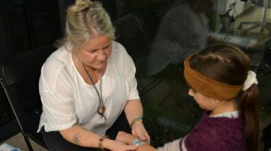 Kobieta siedzi naprzeciwko dziecka i wykonuje na jego dłoni tymczasowy tatuaż. Dziecko podaje rękę, a kobieta precyzyjnie nanosi wzór, trzymając jego dłoń, aby ułatwić wykonanie rysunku.