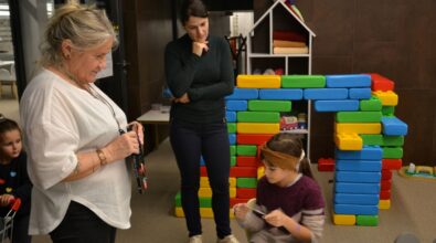 Dziewczynka siedzi przy stole i wykonuje pracę plastyczną z użyciem papieru i drobnych materiałów. Obok stoją dwie kobiety obserwujące jej działania. W tle widoczna jest kolorowa konstrukcja z dużych klocków ułożonych w kształt domu.