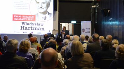 Na zdjęciu widać liczną publiczność zgromadzoną w sali spotkań, siedzącą tyłem do aparatu i skierowaną w stronę prelegenta oraz ekranu z prezentacją. Na ekranie wyświetlony jest portret oraz nazwisko Władysława Hartmana, a całość wskazuje na wykład lub spotkanie historyczne odbywające się w bibliotece.