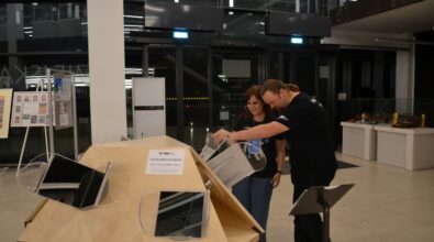 Mężczyzna i kobieta stoją przy drewnianym stanowisku multimedialnym i wspólnie oglądają prezentowane materiały na ekranie. Mężczyzna wskazuje palcem wybrany element, tłumacząc coś towarzyszącej mu osobie. W tle widoczna jest nowoczesna przestrzeń z przeszklonymi ścianami i elementami wyposażenia wystawowego.