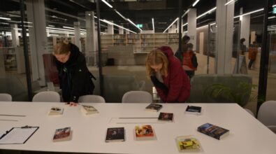 Przy stole dwie osoby przeglądają rozłożone książki i publikacje. Pochylają się nad stołem, oglądając okładki i wybierając interesujące tytuły. W tle widoczna jest przeszklona przestrzeń z regałami bibliotecznymi.