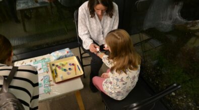 Kobieta siedzi naprzeciwko dziewczynki i wykonuje na jej dłoni tymczasowy tatuaż. Obok na stoliku znajduje się plansza z kolorowymi wzorami oraz drobne akcesoria wykorzystywane podczas zajęć.