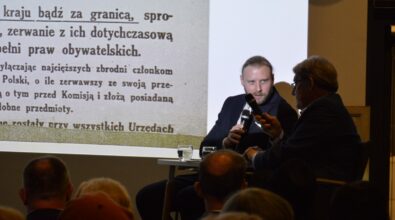 Na zdjęciu widać rozmowę dwóch mężczyzn siedzących na scenie przed zgromadzoną publicznością, z tyłu wyświetlana jest prezentacja z tekstem archiwalnym na ekranie. Uczestnicy wydarzenia słuchają dyskusji prowadzonej w sali spotkań, co wskazuje na debatę lub spotkanie historyczno-edukacyjne odbywające się w bibliotece.