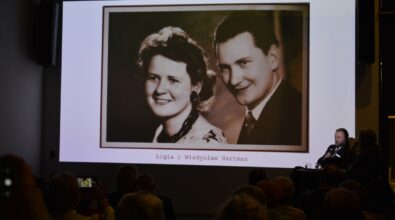 Na zdjęciu widać ekran projekcyjny z archiwalną fotografią przedstawiającą kobietę i mężczyznę, wyświetlaną podczas spotkania w sali wypełnionej publicznością. Uczestnicy siedzą na krzesłach i oglądają prezentację, co wskazuje na wykład lub prelekcję historyczną odbywającą się w bibliotece.