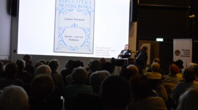 Na zdjęciu widać salę spotkań wypełnioną publicznością, skierowaną w stronę ekranu projekcyjnego oraz prelegenta stojącego obok. Na ekranie wyświetlana jest grafika lub okładka publikacji, a całość przedstawia wykład lub prezentację odbywającą się w przestrzeni bibliotecznej.