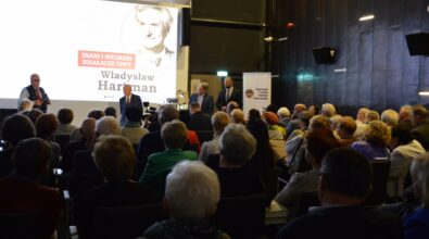 Na zdjęciu widać licznie zgromadzoną publiczność siedzącą na krzesłach w sali spotkań i skierowaną w stronę sceny z prelegentami. W tle widoczny jest ekran z prezentacją poświęconą Władysławowi Hartmanowi oraz roll-up informacyjny, a całość przedstawia wykład lub spotkanie historyczne odbywające się w bibliotece.